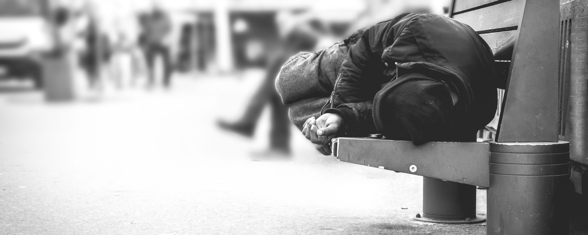 Poor homeless man or refugee sleeping on the wooden bench on the urban street in the city, social documentary concept, selective focus, black and white